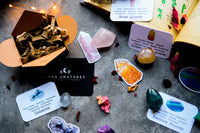 This is a photo of a collection of crystals, books, and cards on a gray surface. The main focus of the image is a black box with “LUNA AMATORES” written on it in gold lettering. The box is open and filled with small pieces of a dark-colored crystal. Surrounding the box are several other crystals, including rose quartz, golden healer quartz, and rainbow fluorite.